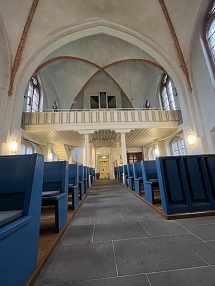Auf dem Bild sieht man einen Teil des Innenraums der Heiligenroder Kirche. Man sieht im Vordergrund blaue Bänke. Oben auf einer Empore befindet sich die Orgel.