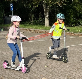 Auf dem Foto sieht man zwei Kinder, die Roller fahren und währenddessen gemeinsam einen Reifen transportieren.